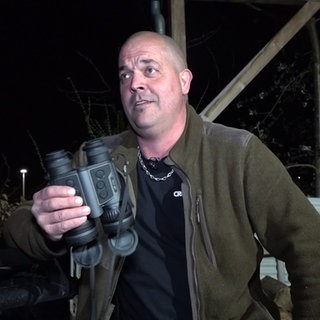 Dennis ist Jäger. Er trägt eine khakifarbene Jacke und hält ein Fernglas in der Hand.