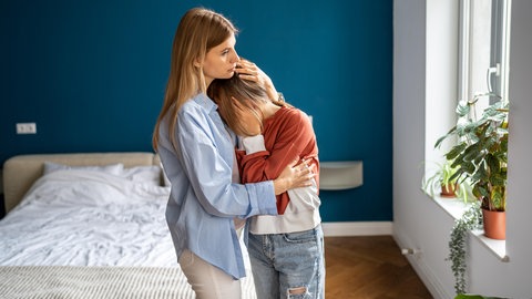 Mutter umarmt tröstend die weinende Tochter zu Hause | Früher Tod eines Elternteils: Wie lassen sich Kinder in ihrer Trauer begleiten?