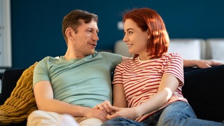 Ein junges Paar sitzt auf dem Sofa und lächelt sich an | Love Languages: So können sie euch helfen, eure Beziehung besser zu verstehen