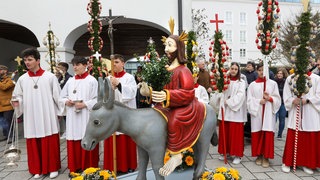 Ministranten stehen an Palmsonntag neben einer Jesusstatue, die auf einem Esel reitet. Für Christen beginnt mit diesem Tag die Karwoche. Am Sonntag vor Ostern, dem Palmsonntag, wird daran erinnert, dass Christus vor seiner Passion triumphal in Jerusalem eingeritten ist, auf einem Esel sitzend. Der Esel war in der semitischen Tradition durchaus ein königliches Reittier. 