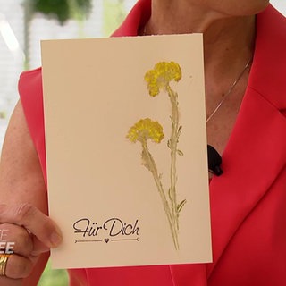 Moderatorin Evelin König hält eine Grußkarte mit einer gedruckten Blüte