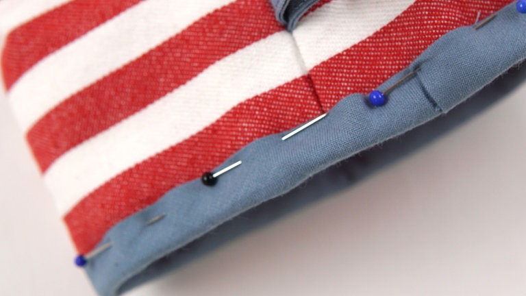 Den Handschuh auf rechts wenden. Das Schrägband über die Kante auf die Außenseite des Handschuhs klappen und so feststecken, dass die erste Naht überdeckt ist.