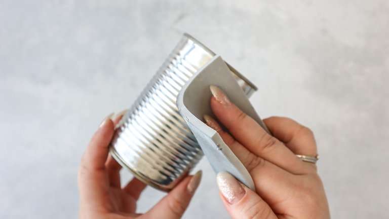 Bei Kleberückständen hilft es, die Dose mit einem feuchten Tuch und etwas Spülmittel zu reinigen. Um die Oberfläche der Dose besser für die Farbe vorzubereiten, wird sie mit feinem Schleifpapier leicht aufgeraut.