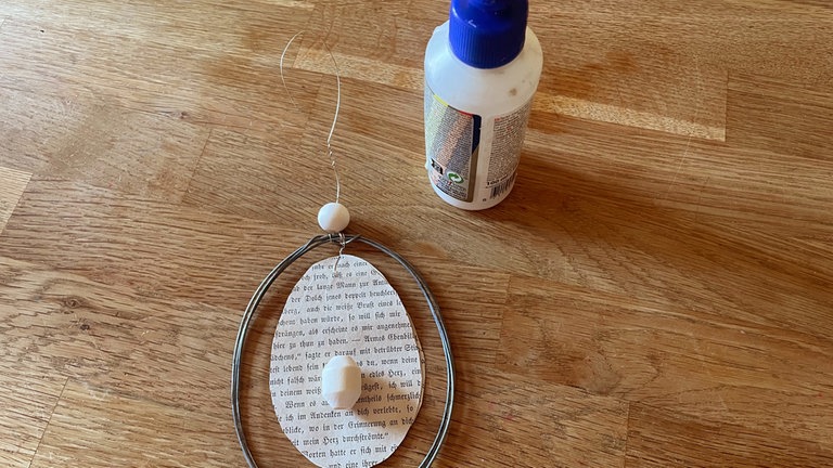 Mit Bastelkleber eine Holzperle im unteren Drittel auf das Papier-Ei aufkleben. Wichtig ist, dass die Bohrung von oben nach unten verläuft.