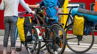 Reisende mit Fahrrädern auf einem Bahnsteig am Bahnhof. Fahrräder sind ein beliebtes Fortbewegungsmittel - im Alltag, aber auch im Urlaub. Wer einen Teil der Strecke mit dem Zug reist, kann es sich leichter machen. Wir erklären, wie.