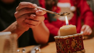 Bleigiessen an Silvester - zwei Hände halten einen Löffel mit Holzstiel, auf dem sich ein Stück Blei befindet, über einer Honigwachskerze