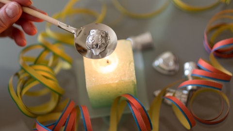 Bleigießen an Silvester über Kerze - eine Hand hält einen Löffel mit Holzstil, auf dem ein Stück Blei in Form einer Krone zum Schmelzen liegt, darunter ist eine angezündete quadratische Kerze umgeben von Luftschlangen