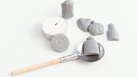 Wachsgießen auf der Silvesterparty - ein Löffel mit Holzstil wie beim Bleigießen liegt auf einem weißen Untergrund, auf ihm ein Stück graues Wachs, auf dem 1000 steht, danneben steht ein angezündetes Teelicht und es liegen mehrere graue Wachsstücke aus einem Wachsgießset daneben
