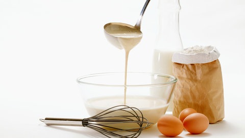 Teiggießen an Silvester anstelle von Bleigießen - in der Mitte steht eine Glasschüssel mit flüssigem Teig, aus einer Metallkelle, die darüber gehalten wird, läuft etwas Teig in die Schüssel, davor liegt ein Metallschneebesen, daneben liegen drei Eier, eine Packung Mehl steht dahinter, genauso wie eine durchsichtige Karaffe mit Milch