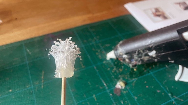 Für das Kleben der Blüte wird zuerst der fransige Streifen als Blütenstempel verwendet. Dafür den Anfang mit Heißkleber an einem Drahtstab oder Holzspieß festkleben. Abschnittweise Heißkleber entlang der geschlossenen Kante auftragen und um den Stab wickeln.