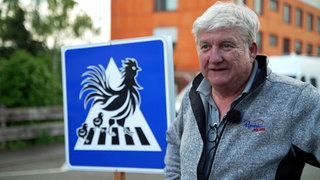 Wolfgang Rombach steht neben einem Straßenschild, auf dem Hühner auf einem Zebrastreifen zu sehen sind.