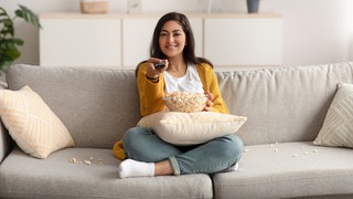 Bereit für den Serien-Marathon: Eine Frau mit Popcorn und Fernbedienung auf dem Sofa