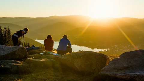 Wandern in Baden-Württemberg - Baden-Württemberg hat tolle Wanderziele: Schwarzwald, Schwäbische Alb, rund um den Bodensee. Diese Tipps sind perfekt für eine Wanderung im Urlaub oder den Ausflug am Wochenende. Das Bild zeigt Wanderer, die auf Felsen sitzen und im Schwarzwald über einen See auf das Panorama im Sonnenuntergang schauen.