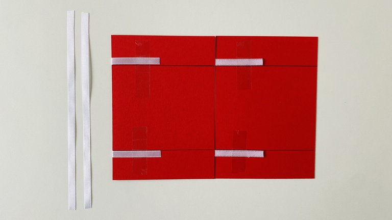 Als nächstes werden die linken Enden der Bänder nach innen umgeklappt und ebenfalls mit einem Klebestreifen fixiert. Auch hier außerhalb der Markierung bleiben. Zwischen den beiden Tonkartonzuschnitten sollte ein kleiner Spalt von ca. 0,5mm verbleiben.