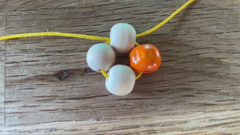 Auf jedes Stück der Kordel nun eine weitere Perle auffädeln. Die vierte Perle auf eine Kordel auffädeln und in die entgegengesetzte Richtung auch die andere Kordel durchziehen. Es entsteht so ein kleiner Ring mit vier Perlen. 