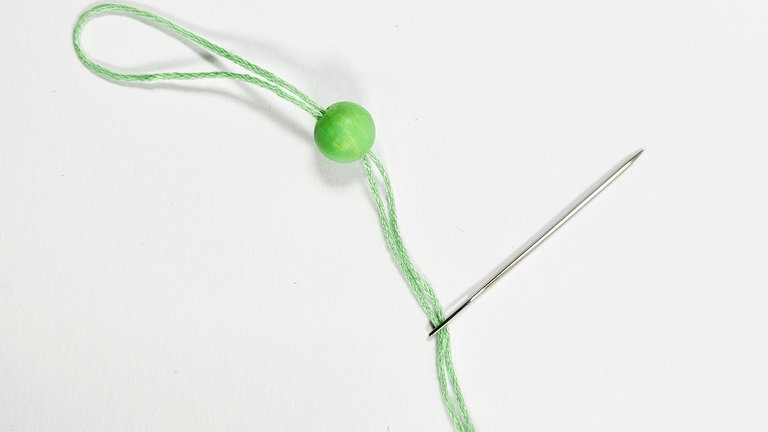 Ein Stück grüne Schnur abschneiden (z.B. Stickgarn) und die beiden Fadenenden durch eine Nadel und durch die grüne Holzperle hindurch ziehen.