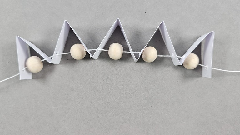 Ein Stück Faden abschneiden, den Faden auf eine Nadel ziehen und nun den Faden unten durch die Löcher hindurch fädeln. Dabei zwischen jedem Blütenblatt eine Holzperle von ca. 8 mm Größe aufziehen. 