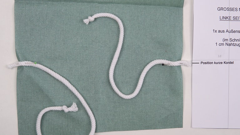 Die Kordel in zwei Teile 37 cm und 30 cm  30 cm und 25 cm schneiden. Jeweils an einem Ende verknoten, um Ausfransen zu verhindern. Die längere Kordel rechts auf rechts und markierungsgemäß so auf dem linken Korpusteil feststecken, dass das unverknotete Ende 1 cm überragt. Die kürzere Kordel ebenfalls rechts auf rechts und markierungsgemäß auf dem Schnittteil platzieren und 1 cm überragend feststecken.