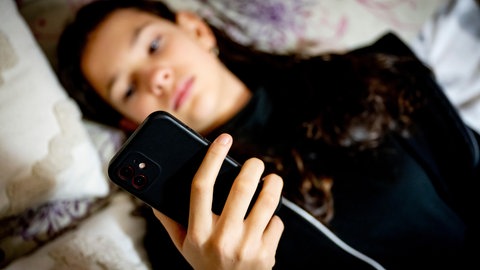 Eine junge Person liegt symbolisch für das von der Wissenschaft festgestellte gestiegene Konsumverhalten digitaler Medien bei Hugendlichen mit dem Smartphone im Bett. Die Forschung zeigt außerdem auch gestiegenes Suchtverhalten während der Pandemie.