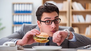 Gestresster Mann ist Burger im Büro und schaut auf die Uhr: Das vegetative Nervensystem ist eng mit Regionen in unserem Gehirn verbunden, die mit Gefühlen, Stress und mit Belastungen zu tun haben. Deshalb führen Stress oder Ärger manchmal zu Verdauungsproblemen. Die Verdauung funktioniert dann nicht mehr richtig.