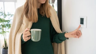 Frau mit Decke um die Schultern und Tasse in der Hand stellt das Thermostat ein: Menschen nehmen Kälte unterschiedlich wahr. Das hat mit dem Körperbau zu tun. Aber das Kälteempfinden hängt auch von äußeren Umständen ab. Die kann man beeinflussen.