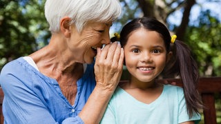Ältere Frau flüstert einem Mädchen etwas ins Ohr.