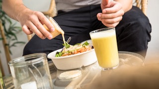 Ein Mann schüttet Soße aus einem Plastikbecher über einen Fertigsalat: Ob Solo Dining ungesund ist, bleibt von Mensch zu Mensch unterschiedlich zu bewerten. Als freiwilliger Trend ist allein essen unproblematisch. Ungewollt oder unter Zwang kann das allerdings ungesund sein.