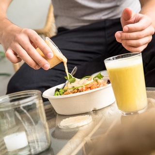 Ein Mann schüttet Soße aus einem Plastikbecher über einen Fertigsalat: Ob Solo Dining ungesund ist, bleibt von Mensch zu Mensch unterschiedlich zu bewerten. Als freiwilliger Trend ist allein essen unproblematisch. Ungewollt oder unter Zwang kann das allerdings ungesund sein.