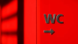 Hinweisschild zur Toilette auf rotem Untergrund: Das Thema Reizblase tritt typischerweise nach der letzten Regelblutung auf – in der Phase, in der der Östrogenspiegel ganz niedrig ist. Von Anneliese Schwenkhagen