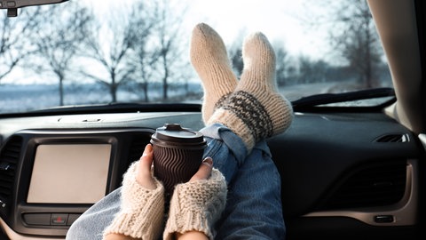 Junge Frau in warmen Socken hat ihre Beine aufs Armaturenbrett eines Autos gelegt und hält einen Becher Kaffee oder Tee: Tee und Kaffee sorgen für Harndrang und kalte Füße. Das liegt am Koffein.