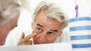 Ein grauhaariger Mann begutachtet vor dem Spiegel im Bad seine Augenringe: Bei Augenringen durch Schlafmangel spielt die dünne Haut unter dem Auge eine Rolle. 