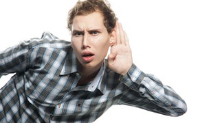 Ein junger Mann hört schlecht und legt die Hand hinters Ohr: In einer lauten Umgebung, einer Bar oder einem Restaurant fällt es auf, wenn man andere nicht mehr gut versteht. Öfter nachfragen oder andere noch mal ansprechen zu müssen sind Anzeichen für eine beginnende Schwerhörigkeit. 