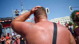 Ein Mann nimmt mit freiem Oberkörper an einer Kundgebung teil. Sein Rücken ist von der Hitze rot und verschwitzt: Unser Körper steht ebenfalls unter großem Stress, um zu verhindern, dass das System überhitzt. Unabhängig von der Außentemperstur muss er seine Temperatur konstant auf ca. 37°C halten. Das ist nötig, damit die lebensnotwendigen Stoffwechselprozesse stattfinden können. Steigt die Körpertemperatur zu sehr, dann zerfallen die körpereigenen Proteine. 