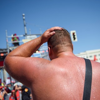 Ein Mann nimmt mit freiem Oberkörper an einer Kundgebung teil. Sein Rücken ist von der Hitze rot und verschwitzt: Unser Körper steht ebenfalls unter großem Stress, um zu verhindern, dass das System überhitzt. Unabhängig von der Außentemperstur muss er seine Temperatur konstant auf ca. 37°C halten. Das ist nötig, damit die lebensnotwendigen Stoffwechselprozesse stattfinden können. Steigt die Körpertemperatur zu sehr, dann zerfallen die körpereigenen Proteine. 