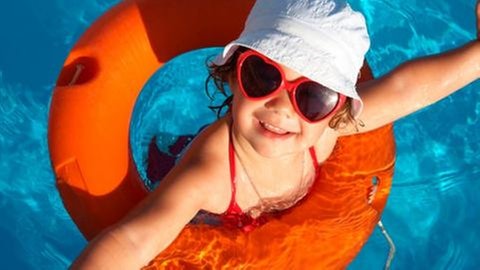 Lachendes Mädchen im Schwimmbad mit Rettungsring, Sonnenbrille und Hut.