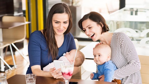 Zwei junge Frauen mit Babys tauschen sich lächelnd in einem Café aus. In Zukunft könnten Ärzte ein Rezept für individuelle Maßnahmen gegen Einsamkeit verschreiben, wie zum Beispiel für ein Treffen für alleinerziehende Mütter.