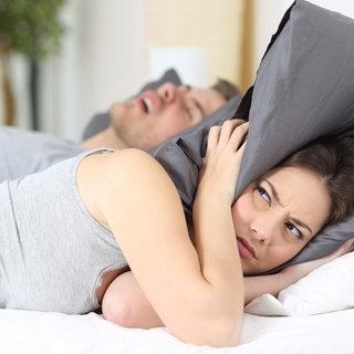 Schnarchen - Frau deckt Ohren mit Kissen zu, während Mann schläft