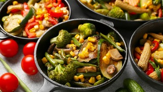 Pfanne mit Zucchini, Brokkoli, Bohnen und Mais. Im Hintergrund sind weitere Pfannen mit Gemüse zu sehen und als Deko liegen neben den Pfannen rohe Tomaten, Bohnen und Gurken.