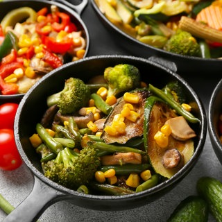 Pfanne mit Zucchini, Brokkoli, Bohnen und Mais. Im Hintergrund sind weitere Pfannen mit Gemüse zu sehen und als Deko liegen neben den Pfannen rohe Tomaten, Bohnen und Gurken.