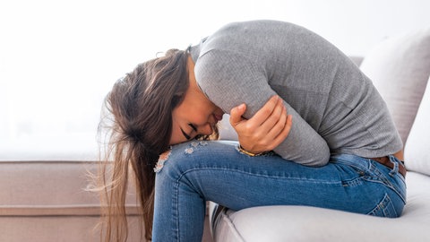 Eine junge Frau auf einer Couch, mit ihrem Oberkörper vorn übergebeugt, schlingt die Armen um ihren Bauch. Chronisch-entzündliche Darmerkrankungen sorgen für mehr als Bauchschmerzen und Durchfall. Betroffene werden in ihrem Alltag und Sozialleben von der Krankheit stark ausgebremst.