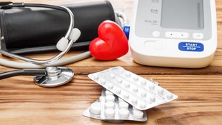 Auf einer Holzunterlage ein rotes Herz, ein Blutdruckmessgerät und blutdrucksenkende Medikamente. Volksleiden Bluthochdruck - falsche Diagnosen, wirksame Medikamente: Bluthochdruck kann falsch behandelt zu Herzinfarkt und Schlaganfall führen. Warum die wahren Ursachen oft unerkannt bleiben, wirksame Medikamente und was Hormone damit zu tun haben.