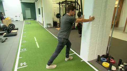 Ein Phisiotherapeut zeigt eine Dehnübung im stehen, die helfen kann einen Krampf in der Wade zu lösen. Dabei lehnt er sich im leichten Ausfallschritt mit dem Oberkörper nach vorne, während er die Ferse des betroffenen Beines auf dem Boden bleibt.