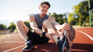 Ein Jogger sitzt auf der Bahn auf dem Boden und hält seine schmerzende Wade fest wegen Muskelkrampf. Sportler nehmen zum Training oft Magnesium, um Muskelkrämpfen und Muskelkater vorzubeugen. Aber neue Studien zeigen: Das bringt nichts, es gibt andere Ursachen für die Krämpfe.