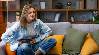 Eine Frau sitzt mit Bauchschmerzen auf einem Sofa und hält ihren Bauch mit den Händen.