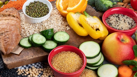 Gesunde Nahrungsmittel wie Gemüse, Vollkornbrot, Hülsenfrüchte, Saaten, Obst auf einem Tisch. Neue Studien zeigen: Unsere Ernährung hat weitreichende Folgen für die Gesundheit – stärker als bislang gedacht. Sie kann chronische Entzündungen, Diabetes oder Herzerkrankungen fördern. Doch eine ausgewogene Ernährung mit Ballaststoffen, Gemüse und Proteinen kann viele Risiken mindern.