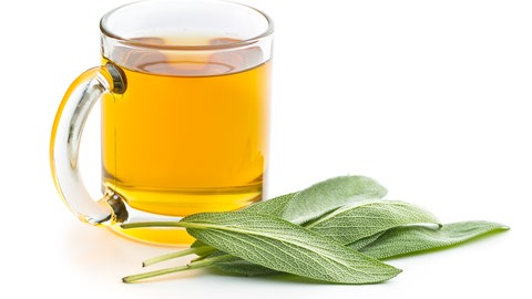 Eine Tasse aus Glas ist mit Salbeitee gefüllt. Vor der Tasse liegen drei Salbeiblätter.