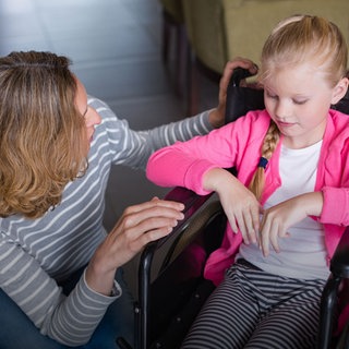 Ein Mädchen in einem Rollstuhl, neben ihr eine Frau. Bei Hilfsmitteln wie Prothesen, Windeln, Rollstühlen wollen Krankenkassen häufig die Kosten zunächst nicht übernehmen.