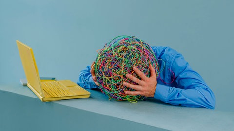 Symbolbild für ADHS wie es in der Wissenschaft und auf Social Media diskutiert wird: Person sitzt vor einem Laptop und hält den Kopf in den Händen, wobei der Kopf durch ein buntes Knäuel aus Gummibändern dargestellt wird