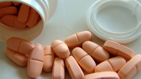 Symbolbild für die medikamentöse Behandlung von ADHS, während die Wissenschaft auch nicht-medikamentöse Therapieansätze für die Erkrankung erforscht: Orangefarbene Tabletten liegen neben einer weißen Medikamentendose auf hellem Untergrund 
