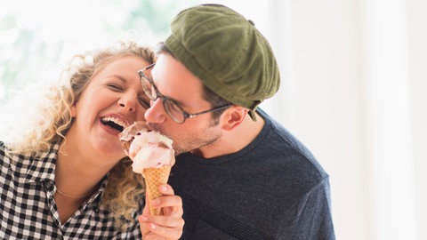 Ein Paar zusammen beim Eis essen. Der Mann beißt in die Eiscreme und die Frau lacht herzlich darüber.